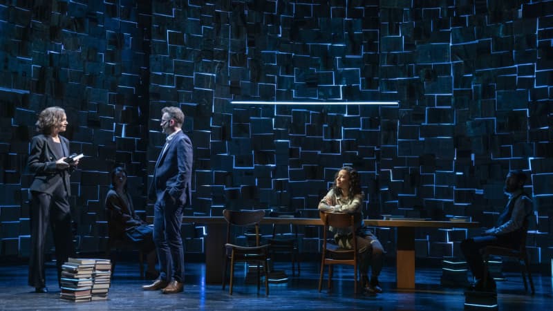 The background is a large wall constructed of open books, and blue light shines through the cracks. Two actors sit on opposite sides of a table, one crouches in a chair, and two talk to the side.