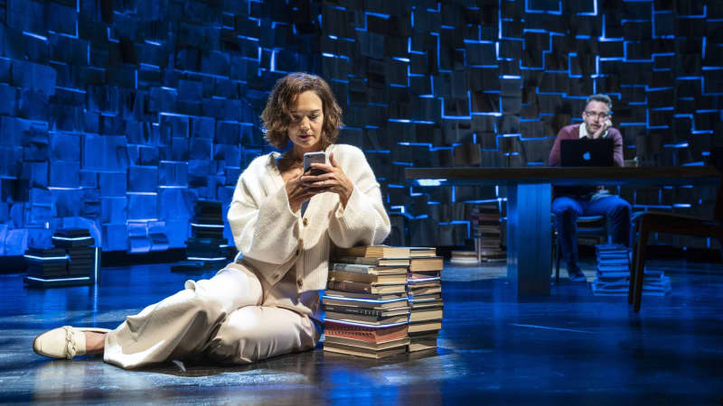 A woman sits on the floor with her legs to the side, leaning an elbow on a stack of books as she types on her cell. Behind her, a man sits at a table with a laptop and holds his head in his hands.