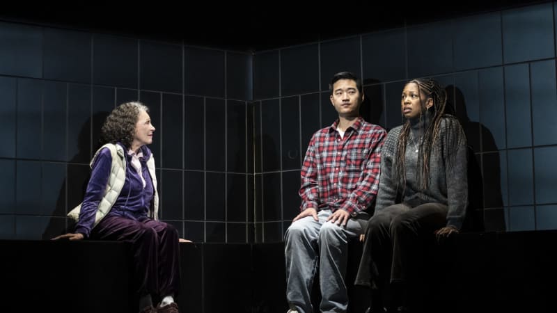 Three people sit on a black L-shaped bench. On one side is a woman with grown-out grey roots. A younger man and woman sit next to each other on the other side.