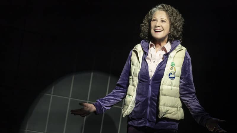 A spotlight on a woman with brown curly hair and grown-out silver roots speaking to the audience. She wears a purple velvet tracksuit underneath a beige vest with a round "Juror" sticker on it.