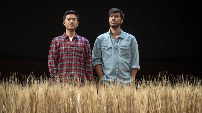 Two men stand side-by-side in a field of wheat with their hands in their pockets. They both have short, dark hair and are wearing button down shirts, one is red plaid and the other is a light denim.