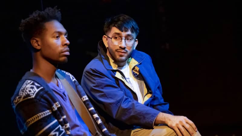 Two actors sit next to each other, one staring straight ahead and one staring at the other person. One wears glasses, a blue jacket, and khakis. The other wears a navy cardigan over a purple tee.