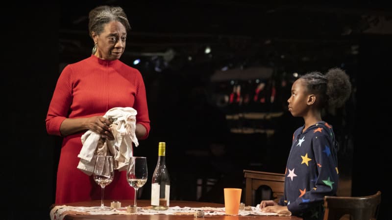 A grandmother and young granddaughter stand at a dinner table. The grandmother has a dish towel in her hand and wears a red sweater dress. The granddaughter is in footie PJs.