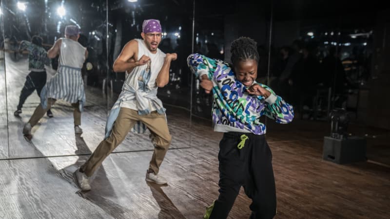 A dance teacher and young student enthusiastically hip-hop dance. They're both dressed in loungewear. They're in a room with mirrored walls and wooden floors.