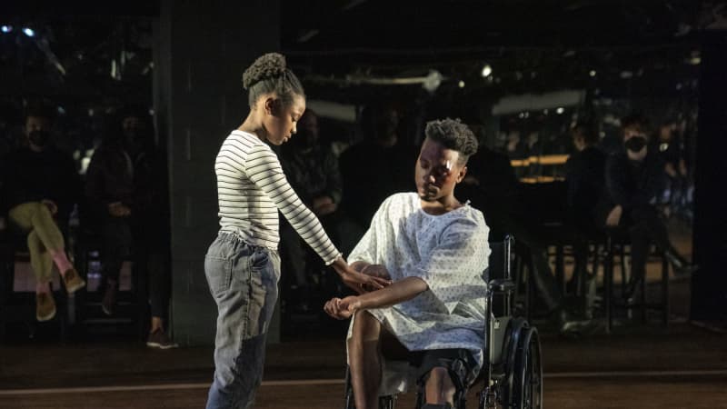 A man in a hospital gown sits in a wheelchair. There is a bandaid above his bruised eye. His young daughter stands by his side and gently touches the scars on his arm.