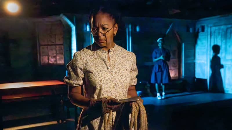 A dark skinned woman dressed in clothes from the 30s holding a rope and a letter looking off stage. She is lit in dark dramatic lighting. A girl stands in the distance behind her. 