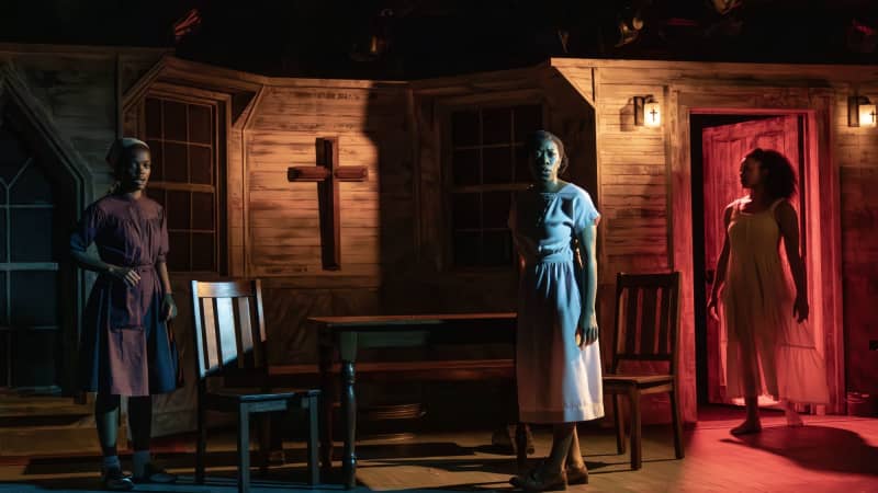 Three dark skinned women standing in a dark lit kitchen. One woman in a white dress is walking in a door that is lit with bright red light. 