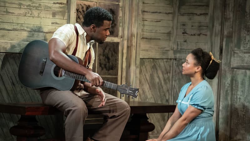 A man sits with an acoustic guitar on a church bench looking at a woman kneeling in a blue dress and looking at him. 