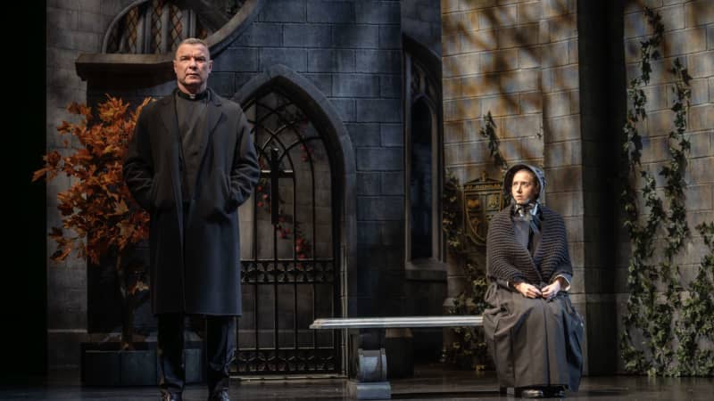 A man is standing at the end of the stage with a long black coat on looking out while a nun is sitting on a stone bench behind him looking at him. 