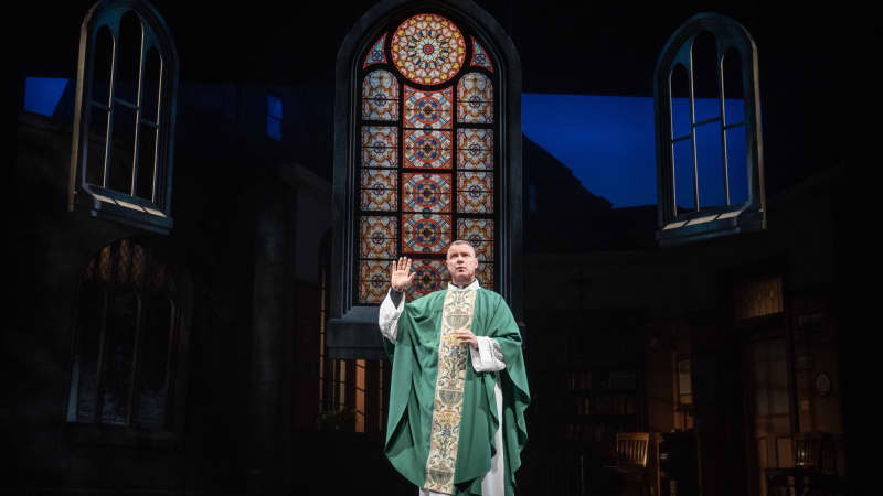 A priest on a green robe is standing with is hand up in front of stained glass windows. 