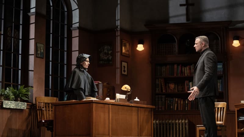 A nun standing behind a desk staring intensely at a priest who has his arms out pleading his case. 