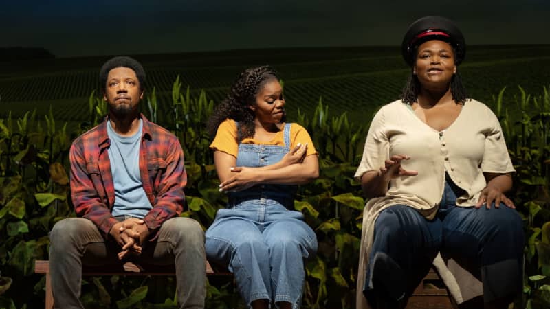 A Black woman in a bus driver's hat sits on a block and mimes driving. A Black man and woman sit on a bench like passengers, the woman mimes rocking a baby. A field of crops is behind them.