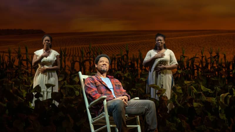 A smiling Black man in a flannel sits in a rocking chair and stares nostalgically into the distance. Behind him, two Black women stand amid crops and each hold a hand over their chest and belly.