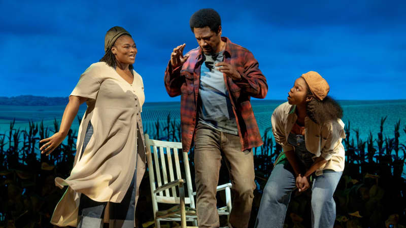 Two Black women wearing backwards hats taunt and tease a Black, flustered man wearing a flannel. They stand on a wooden platform with a rocking chair on it in front of a field of crops at dusk.