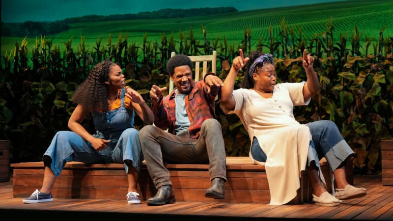 Three Black actors on stage, one man between two women, sit on the edge of a wooden platform in front of a tobacco field. They make animated gestures and laugh together. They wear 60s rural clothing.