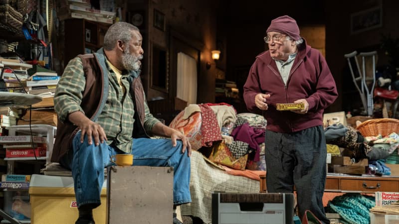 Two old men, on Black and one white, have a conversation in a messy living room. The Black man sits on a pile of stuff while the white man stands and holds a trinket box.