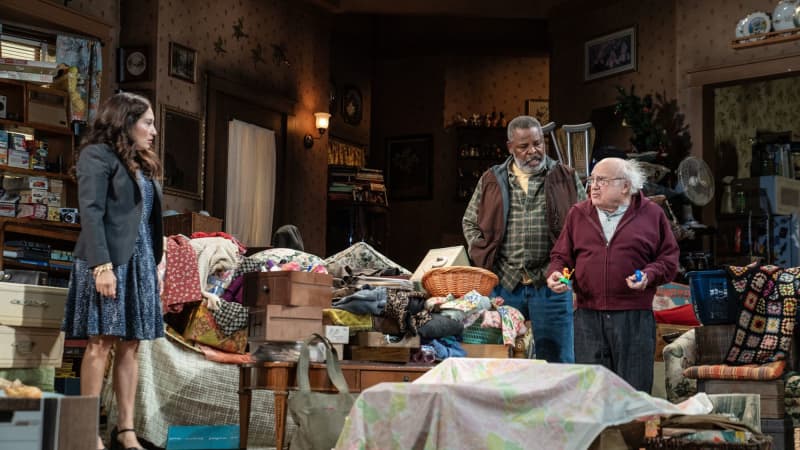 A woman stands on the opposite side of a messy living room, listening to an old man speak while his best friend stands behind him.