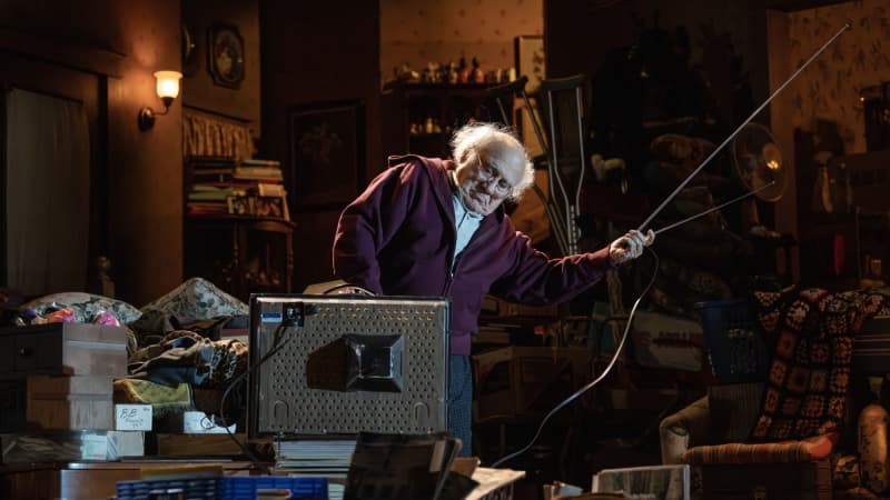 An old man in a zip up, lounge pants, and glasses fusses with the antennas on an old TV. His home is overcrowded with items.