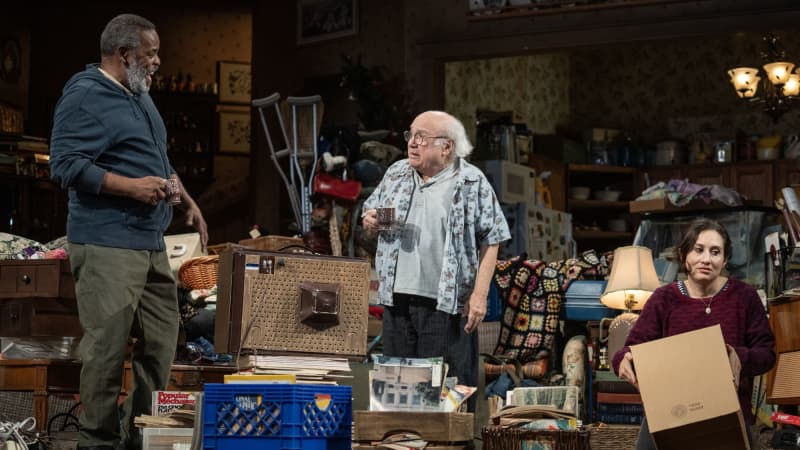 Three actors on stage on a set dressed like an extremely cluttered home. Two men laugh and chat while an annoyed woman packs a box.