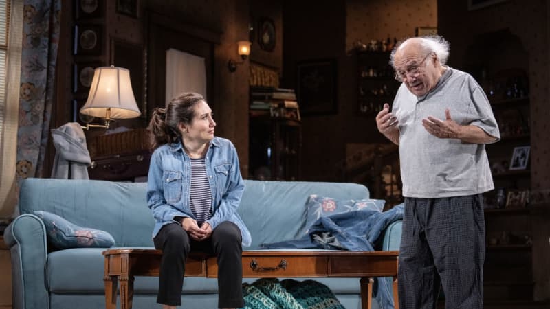 A woman sits on a clean coffee table in front of a clean couch while her father stands next to her and speaks.