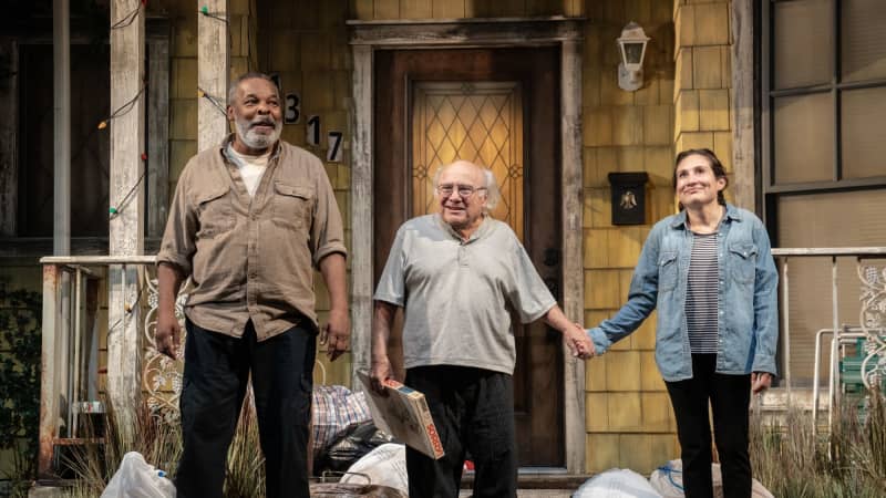 At the curtain call, three smiling actors stand in front of a yellow house surrounded by trash bags. Danny DeVito holds a SORRY! game box in one hand and holds Lucy's hand in the other.