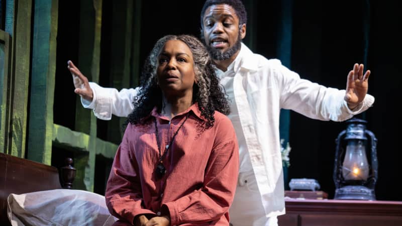 A Black woman with graying hair sits on the edge of a bed while a Black man with graying hair stands behind her, dressed in white and arms outstretched.