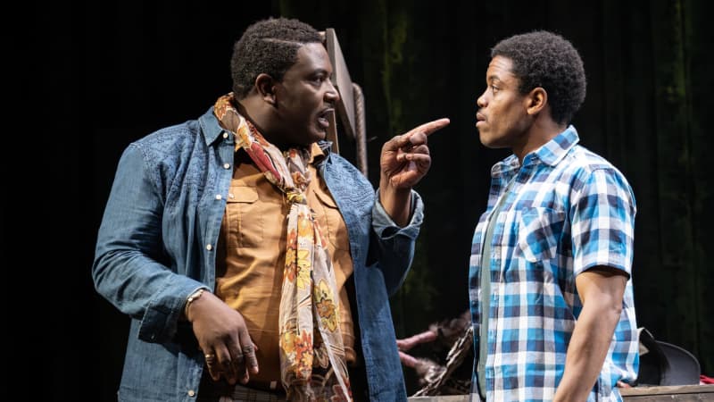 A Black man wearing a florla scarf with a denim jacket points his finger at a younger Black man wearing a blue flannel.