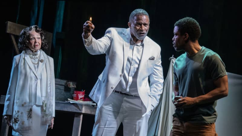 An old Black man and woman dressed in white clothes stand across from a young Black man in a t-shirt and pants. The old man holds a golden lighter with a tall flame.