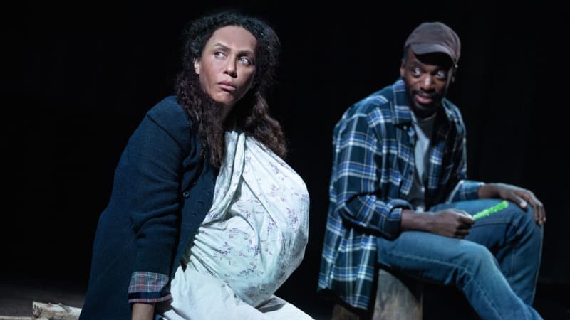 A woman, covered in dirt, with a baby strapped to her chest holds a hammer and stares cautiously to the side while a man in a flannel and jeans sits next to her on a bucket with candy in his hand.