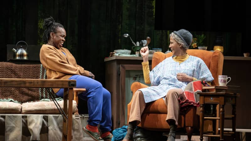 A young Black woman sits across from and laughs with an old Black woman sitting in an orange recliner chair.