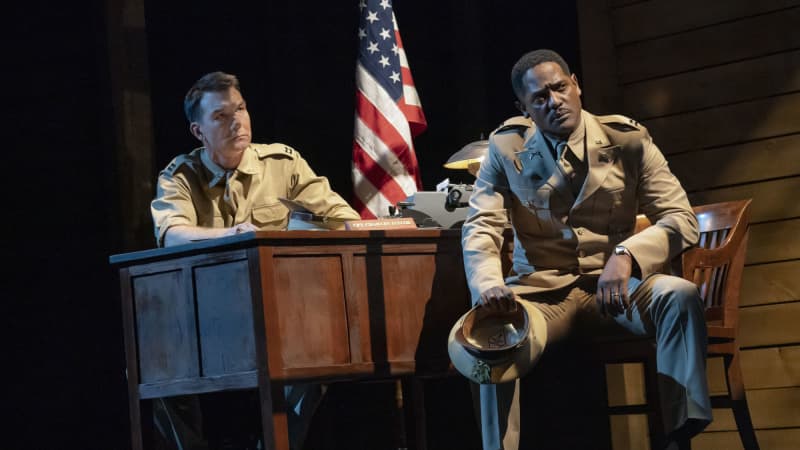 A white solider sits behind a wooden desk, an American flag pole in the corner of his office. A Black soldier sits away from the table with concern and holds his hat on his knee.