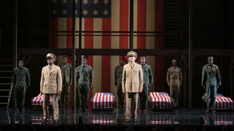 A group of men in soldier's uniforms stand at attention in front of and behind a row of cots, each one with an American flag laying over it. A large American flag is projected in the background.