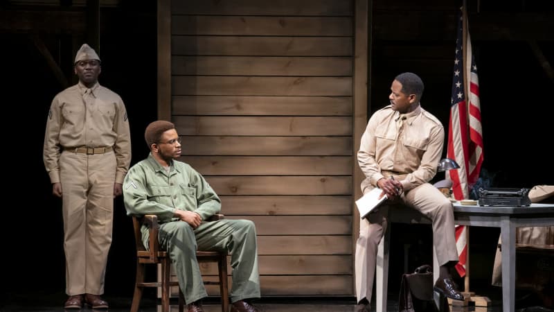 A Black soldier in a tan uniform sits on the edge of an office desk and speaks to a younger Black man with glasses in a green uniform. Another soldier in a tan uniform stands at attention behind them.