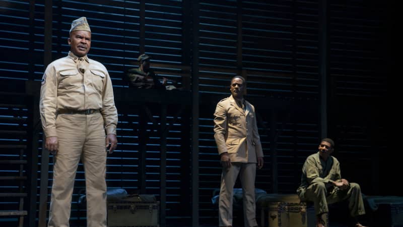 A Black soldier in a tan uniform and garrison cap stands facing the audience, while two other soldiers watch him with concern.