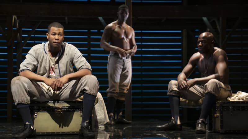 Two Black actors sit on trunks while another stands behind them. Two are shirtless and wear the bottom half of their uniforms. One is in sweats and is more brightly lit by the stage lights.
