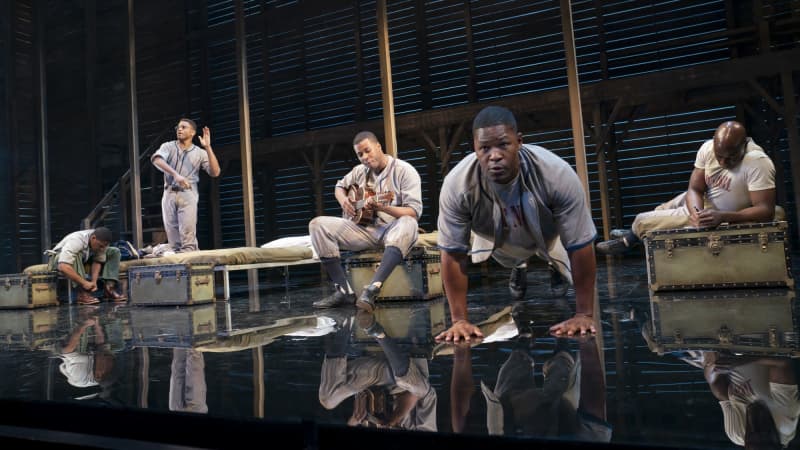 A group of Black men hang around their cots and trunks. One ties his shoe, one is doing push-ups, another is laying down, and one is playing guitar.