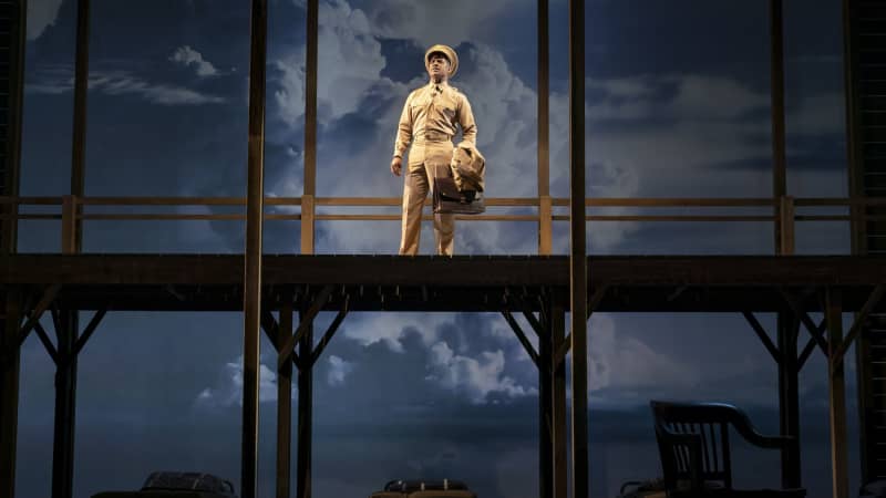 One actor dressed in a soldier's uniform holding a briefcase stands on a high platform and looks out into the distance. A blue cloudy sky is behind him.