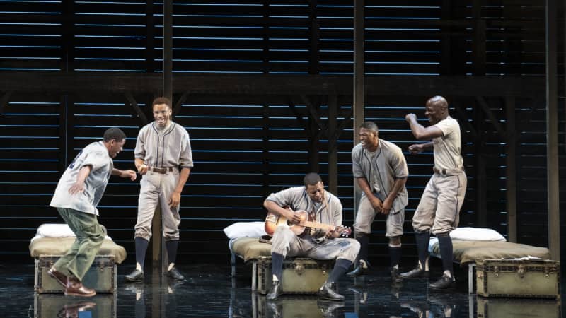 A group of Black men smile and dance around their cots and trunks. One sits on a trunk playing the guitar.