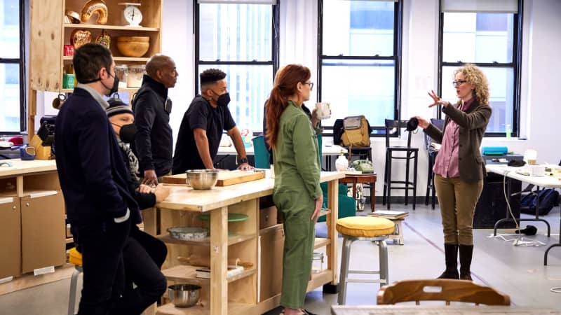 A group of actors and the playwright stand and lean around a kitchen island set while the director speaks to them.