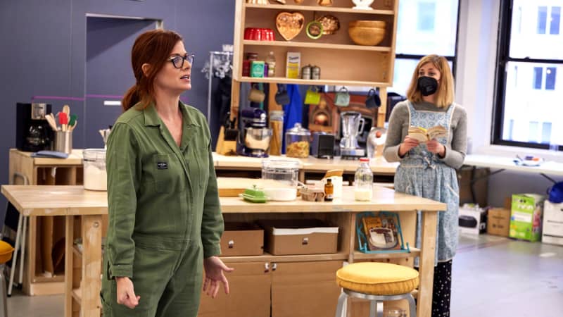 A white woman with auburn hair and glasses stands among a kitchen rehearsal set. She wears a collared green jumpsuit. Another woman stand behind her at an island counter.