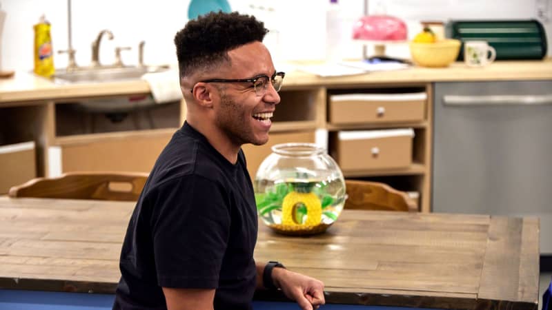 A Black man wearing glasses laughs while sitting at a wooden table in a rehearsal set that looks like a kitchen. 