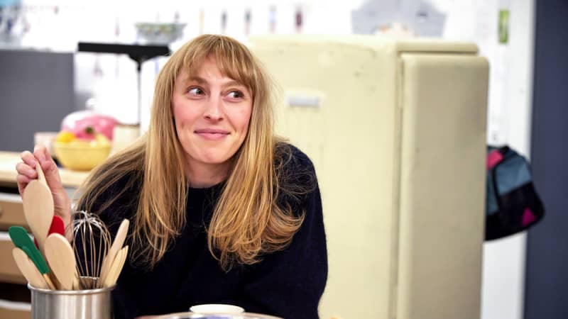 A white woman with blonde hair and bangs leans on the kitchen counter and fiddles with a can full of baking utensils.