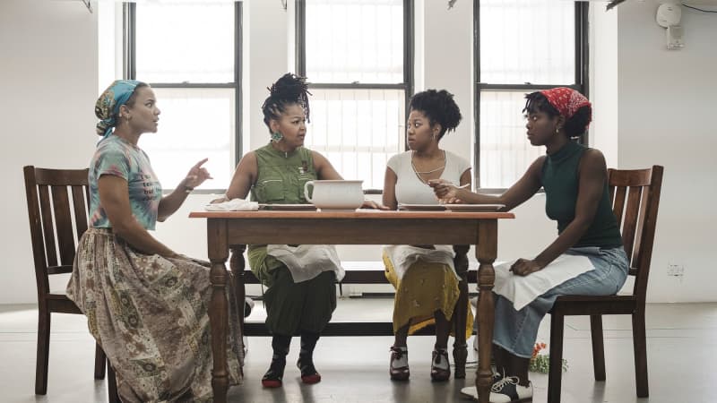 Three young Black women and one older African American woman sit around a dinner table in the rehearsal studio and appear to be in a dramatic conversation.