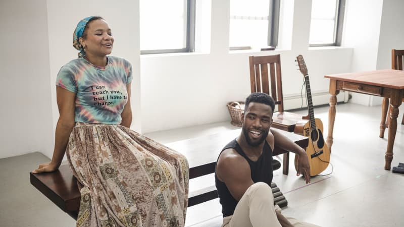 A young black woman sits on a bench in rehearsal while a young black man leans against the bench on the ground, his guitar in the background.