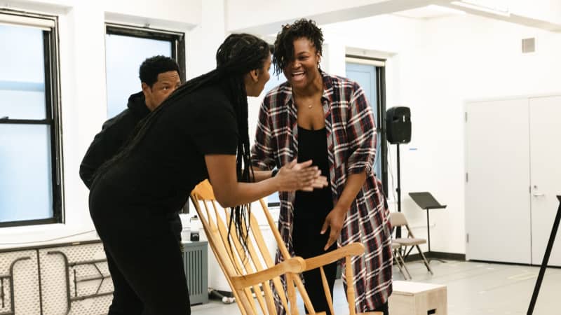 The cast of Home on a small wooden platform in a bright rehearsal room all standing around a rocking chair laughing and smiling. 