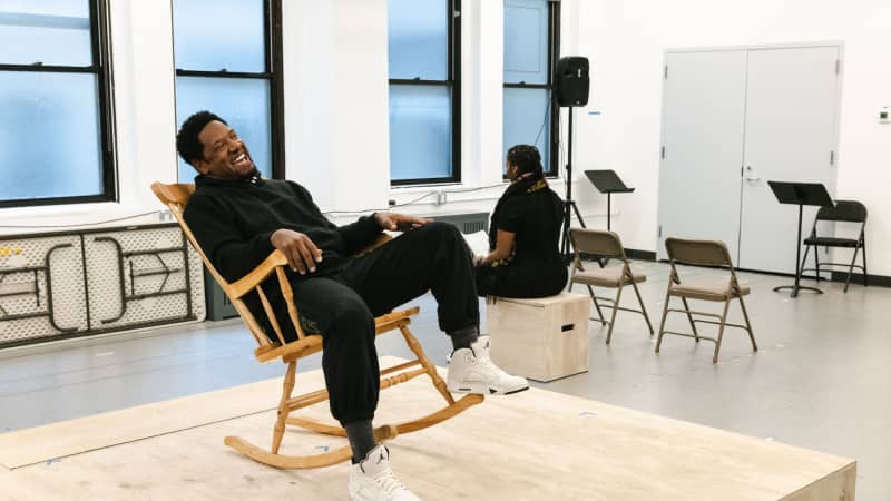 Tory Kittles, a dark skinned man, sitting in a rocking chair on a small platform in the middle of a bright rehearsal room. 