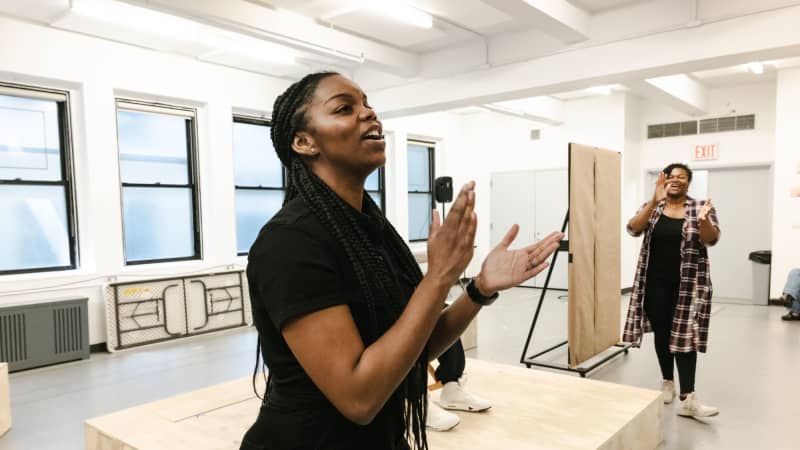 Brittany Inge is standing in the front of a bright rehearsal room and is clapping and singing.