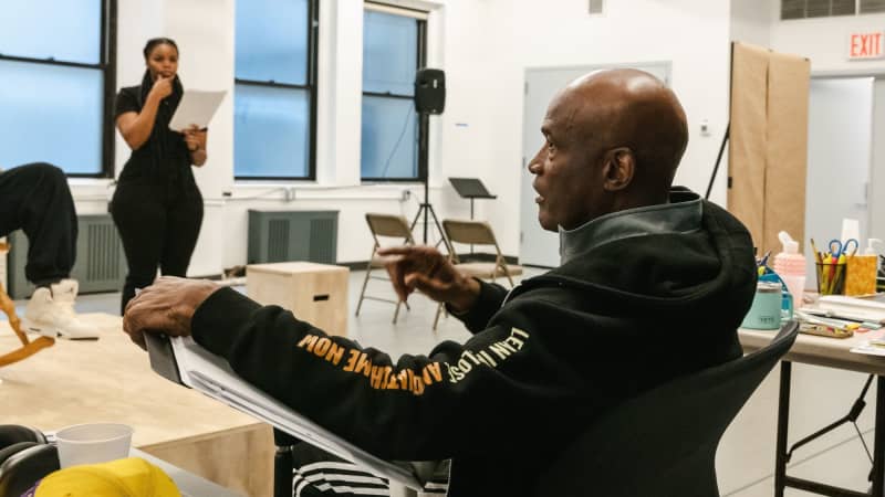 Kenny Leon a dark skinned black man sitting at a table with papers directing a cast in a brightly lit rehearsal room. 