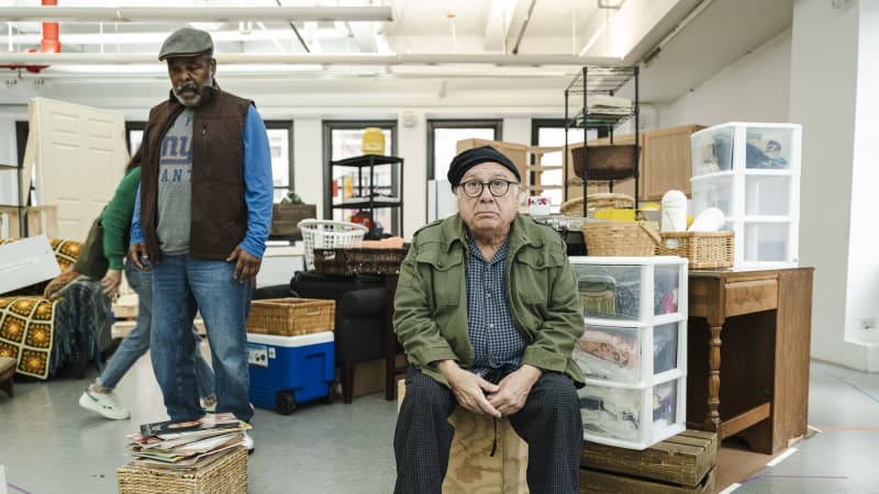 Danny DeVito sits on a wooden box among a rehearsal set decorated like a messy house. He and Ray Anthony Thomas, standing behind him, share a sad face.