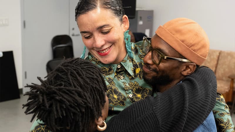 Director Patricia McGregor and actors Ngozi Anyanwu and Daniel J. Watts wrap their arms together in a tight group hug during rehearsal.